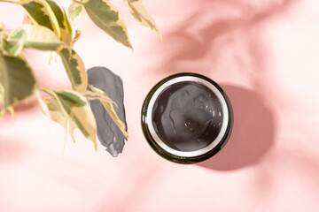 Cosmetic clay mask in an open jar and smeared on a pink background with green leaves. A purifying...