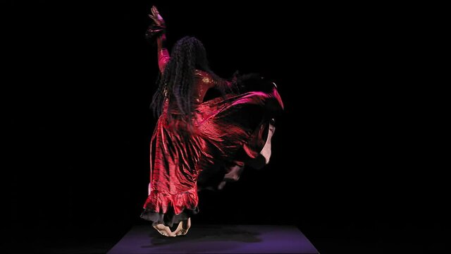 Barefoot gypsy woman with long hair in a red suit is dancing on a black background. Overall plan