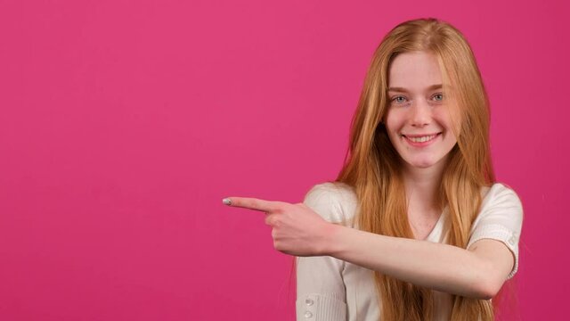 Close up of a confident looking red haired woman with freckles that is pointing to the leftwith finger and smile, standing isolated over pink background in studio.
