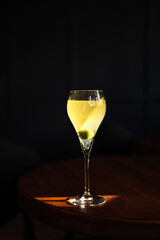 A sweet and sour refreshing french 75 sparkling gin cocktail in a flute glass, olive garnish, in direct hard sunlight with long shadows, on a wooden table, vertical photo with copy space