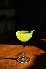 A sweet and sour refreshing basil gin sour cocktail in a coupe glass in direct hard sunlight with long shadows, on a wooden table, vertical photo with copy space