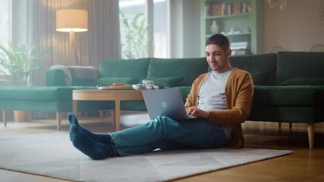 Handsome Adult Man Using Laptop Computer, while Sitting on Living Room Floor in Cozy Stylish Apartment. Attractive Man is Online Shopping on Internet, Watching Funny Videos on Streaming Service.