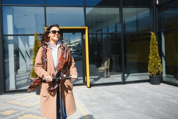 Young beautiful woman in a jacket smiles and rides an electric scooter to work along office buildings