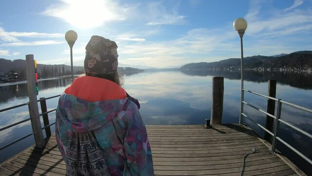 A woman in a flower beanie walking on the pier along Woerthersee Lake in Austria, and enjoying the sunlight. The lake is surrounded by high mountains. Small villages at the shore. Calmness
