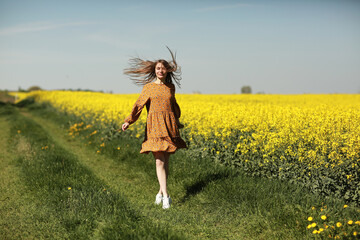 beautiful young woman walks in a field of yellow rapeseed. Girl brunette long hair fly in wind dressed in dress and straw hat. summer holiday concept