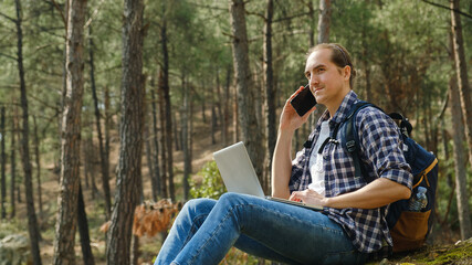 Obraz premium Young man sitting on ground at forest talking on smartphone and working on laptop