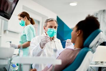 Smiling dentist with face mask talking to Black woman during dental procedure at dental clinic. - obrazy, fototapety, plakaty