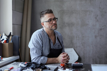 Linocut master in apron or craftsman sitting at workplace thinking about new project. Bearded...