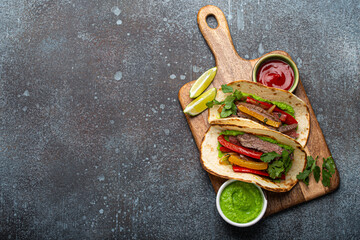 Traditional Mexican dish Beef fajitas tacos served on wooden cutting board with tomato salsa and guacamole on rustic stone background from above with space for text, American Mexican food 