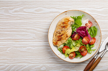 Grilled chicken breast fillet with vegetables salad of fresh tomatoes and salad leaves on white plate on wooden background from above with space for text, healthy dinner or diet meal 