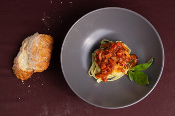 Top view of dish or gray plate with pasta or fettuccine with red or tomato sauce, bacon, pepperoni or calabresa and basil, next to a piece of bread