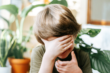 Portrait of child Kid boy hiding face in green t shirt in living room at sunny day. Stylish interior with a lot of plants. Home gardening, love of houseplants. Spring time. 