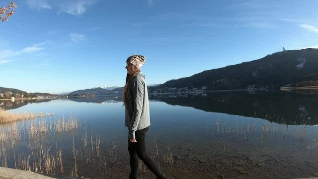 A woman in a flower beanie walking on the pier along Woerthersee Lake in Austria, and enjoying the sunlight. The lake is surrounded by high mountains. Small villages at the shore. Calmness

