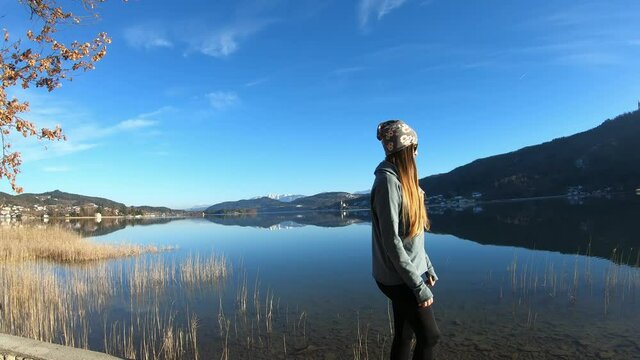 A woman in a flower beanie walking on the pier along Woerthersee Lake in Austria, and enjoying the sunlight. The lake is surrounded by high mountains. Small villages at the shore. Calmness
