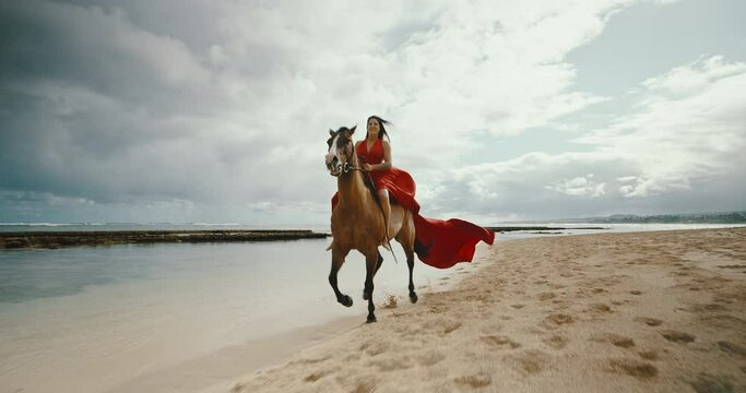 Beautiful woman horseback riding on the beach in flowing red dress, cinematic slow motion