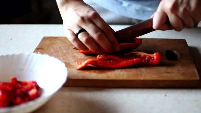 woman cuts red bell pepper into strips on a wooden cutting board with a large sharp knife. Concept of cooking at home.