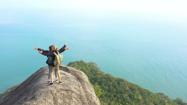 Drone shot of young caucasian man and woman stand on top of the mountain. Couple backpacker celebrate with arms raised and look around at view point. Aerial shot, Nature, Travel and Adventure concept.