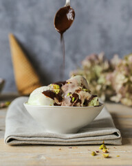 Pistachio vanilla chocolate ice cream garnished with chocolate sauce, sprinkled with pistachio nuts in a bowl on a wooden table.