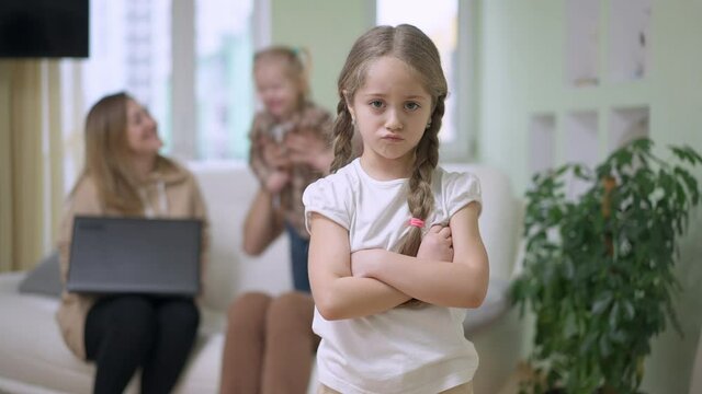 Sad pretty little girl crossing hands looking at camera with dissatisfied facial expression as parents playing with baby sister at background. Upset child posing at home. Envy and jealousy concept