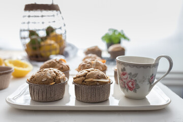 Two just baked homemade muffins and a beautiful cup of coffee in the foreground accompanied by more blurred muffins over a white ceramic tray.