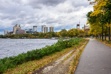 Fototapeta na wymiar Deserted riverside paved path lined with autumn trees in a public park under grey stormy sky. High rise office and residential buildings are visible in background. Boston, MA, USA.
