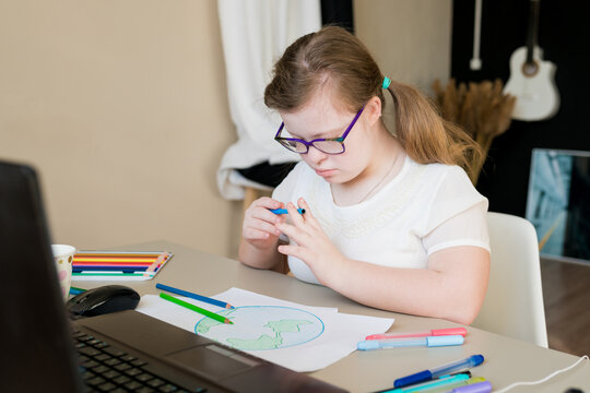 Teenager girl with Down syndrome drawing planet Earth for online lessons. Distance learning by video call on laptop. E-learning and creative development for kids with special needs concept