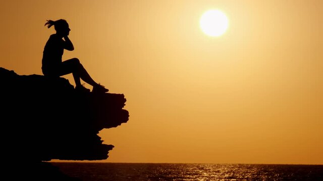 Silhouette of hipster woman puts headphones and enjoys music while sitting on cliff face looking at sea at sunset.