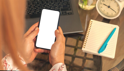 Woman using smartphone with blank whites screen, feminine mockup concept with notebook and alarm clock on the background