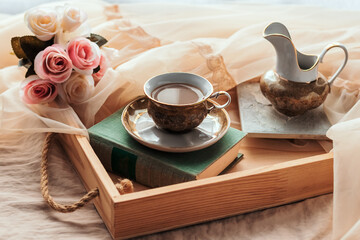 Coffee in bed, romantic morning. A cup of coffee, a book and a bouquet of flowers on a wooden tray on a silk bed. Home cozy interior, lifestyle.	