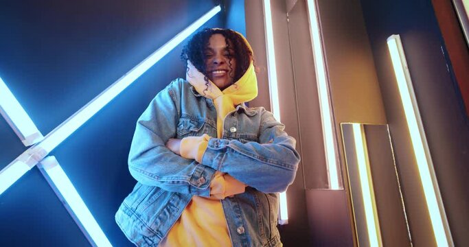 Handsome black woman dancing standing by colorful striped lights wall in night disco club. African American urban female wearing yellow hoodie jeans jacket moves rhythmically to music beat