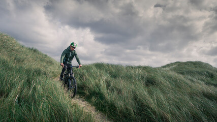 Man with bike in sand dunes with grass at coast in Denmark, dark clouds in dramatic sky.