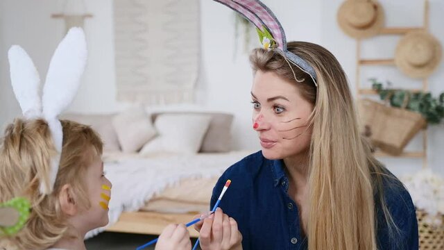 Beautiful happy son and mother in headbands with bunny ears decorating eggs using paintbrush and bright colors of paint, preparing to celebrate Easter holiday, having fun spending time together at hom