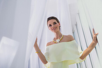 Portrait of a young brunette girl model in a pale lemon dress. Jewelry. Standing by the window.