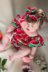 Little toddler girl in a colorful dress and a headscarf on her head. Sitting on a bed with flowers.