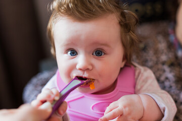 Portrait of a little blond toddler girl. Spoon eating. Dirty mouth. Mom feeds the baby.