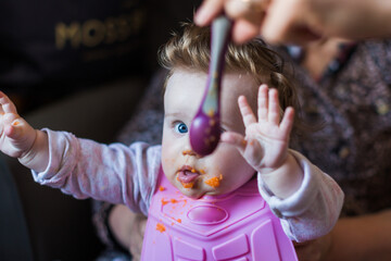Portrait of a little blond toddler girl. Spoon eating. Dirty mouth. Mom feeds the baby.