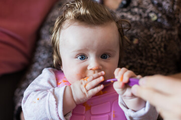 Portrait of a little blond toddler girl. Spoon eating. Dirty mouth. Mom feeds the baby.