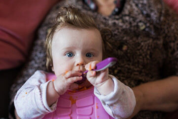 Portrait of a little blond toddler girl. Spoon eating. Dirty mouth. Mom feeds the baby.