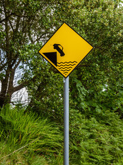 A yellow street sign tells people to beware of their car falling off the cliff into the water.