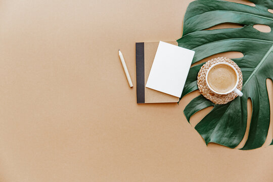 A simple trendy office Desk. Coffee cup, monstera leaf, blank card and notebook .Home Office desktop with space copying. Top view, copy space. Minimal concept