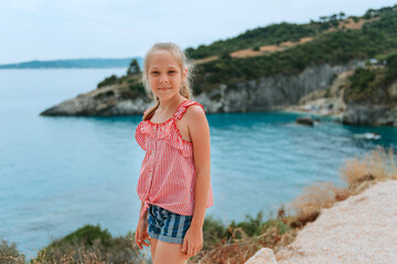 little girl stands on the sea coast and smiles. Adorable little girl child looks at the flower on beach. Portrait of a child with wet long blonde hair in dress sniffing the fragrance of wild purple