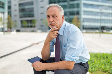 Businessman using his tablet outdoor while sitting on a bench in front of his office