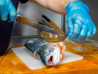 The gloved hands of the cook cut off the fins on the salmon carcass with a knife. Cutting fresh fish in the kitchen on a cutting board.