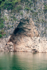 Guandukou, Hubei, China - May 7, 2010: Wu Gorge on Yangtze River. Closeup of huge cave on shoreline of green water set in gray-brown rock cliff. Hanging green tree foliage and plants. 