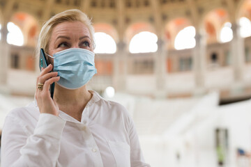Stylish mature lady in protective face mask speaking on smartphone in art museum