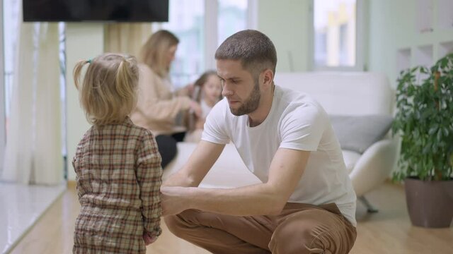 Concentrated young father putting dress on baby daughter with blurred mother braiding girl hair at background. Caucasian parents taking care of children at home in living room. Family concept