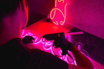 Back view of man with glue gun making modern neon signboard in dark workshop 