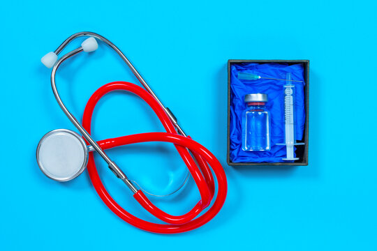 Stethoscope to make medical examination and box with syringe and glass vial of vaccine for new virus on blue background, top view, copy space for advertising text.