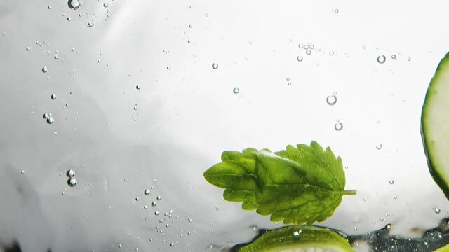 Person throws slices of cucumber lime and mint leaves into clear water on white background macro bottom view slow motion