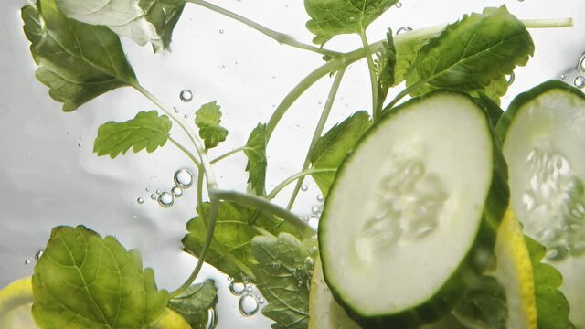 Fresh mint and slices of juicy cucumber and lemon fall into water on white background extreme close bottom view slow motion